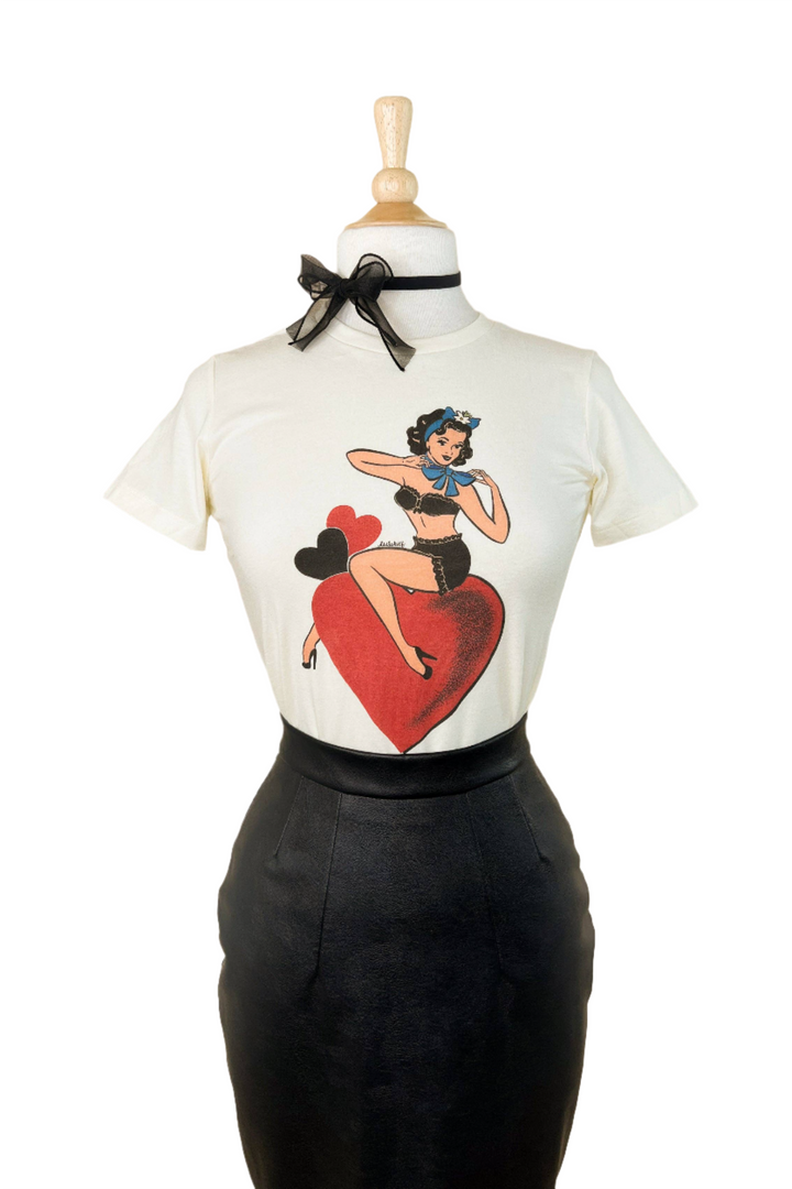 A fitted tee in ivory color with a graphic print of a character sitting on a heart, displayed on a mannequin paired with a black skirt.