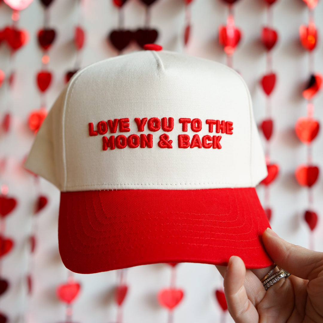 White and red cap with 'LOVE YOU TO THE MOON & BACK' text held in front of a heart-patterned background.