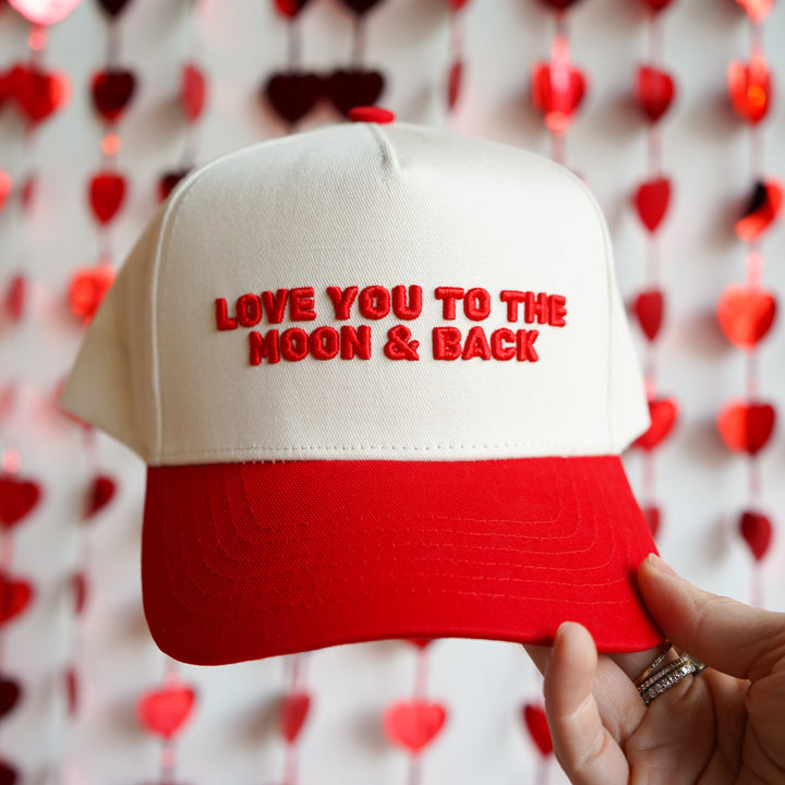 White and red cap with 'LOVE YOU TO THE MOON & BACK' text held in front of a heart-patterned background.