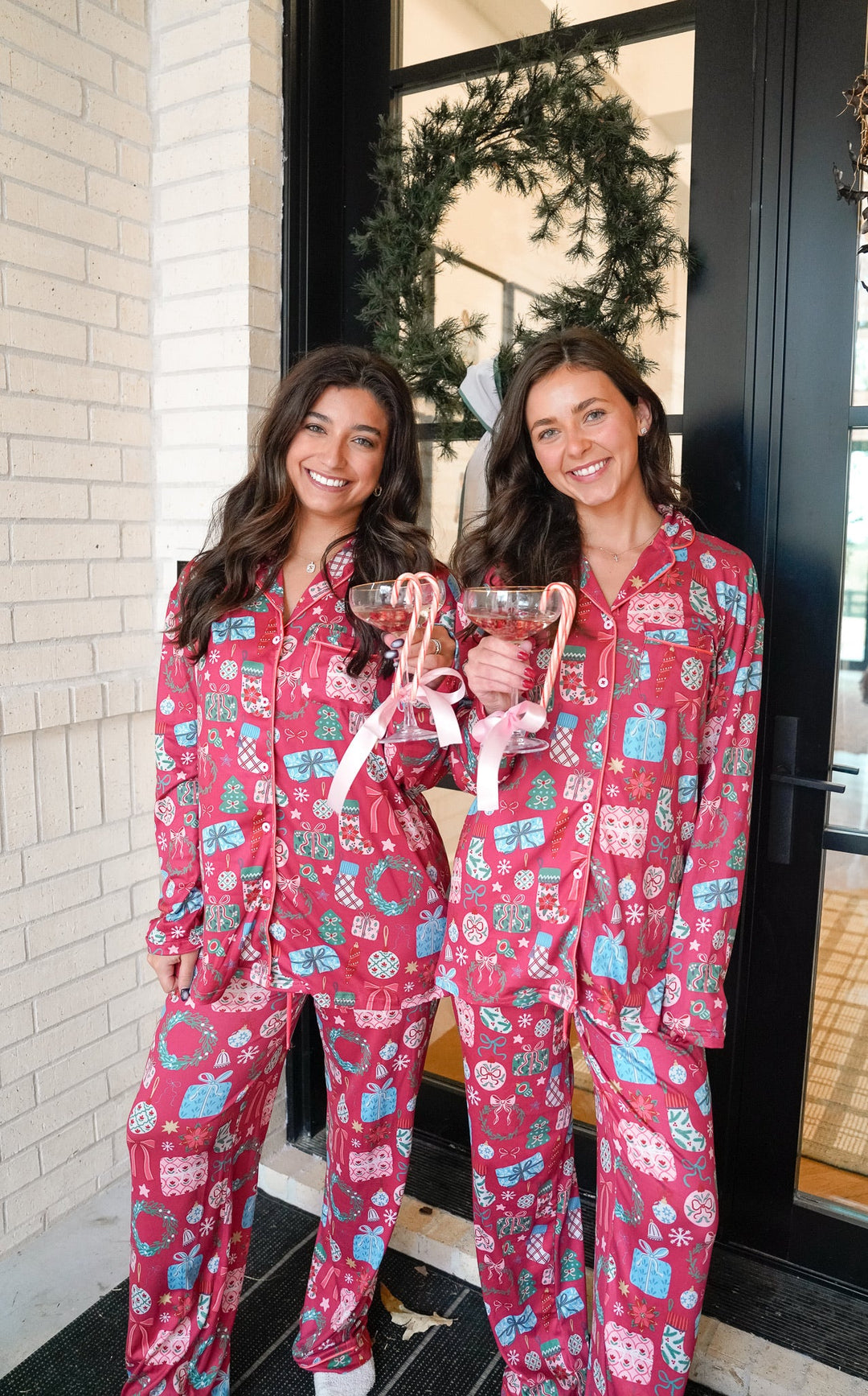 Two women wearing pink pajamas with a festive pattern, standing in front of a door with a wreath.