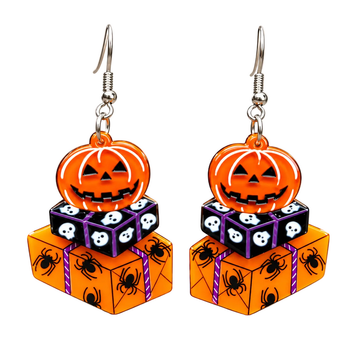 A pair of Halloween-themed dangle earrings featuring orange jack-o'-lantern charms, stacked gift boxes with spider and skull motifs, and silver fishhook wires, displayed against a softly lit neutral background.