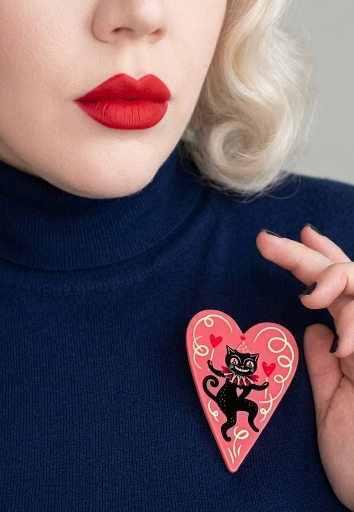 Person wearing a black turtleneck with a pink heart-shaped brooch featuring a black cat design by Johanna parker