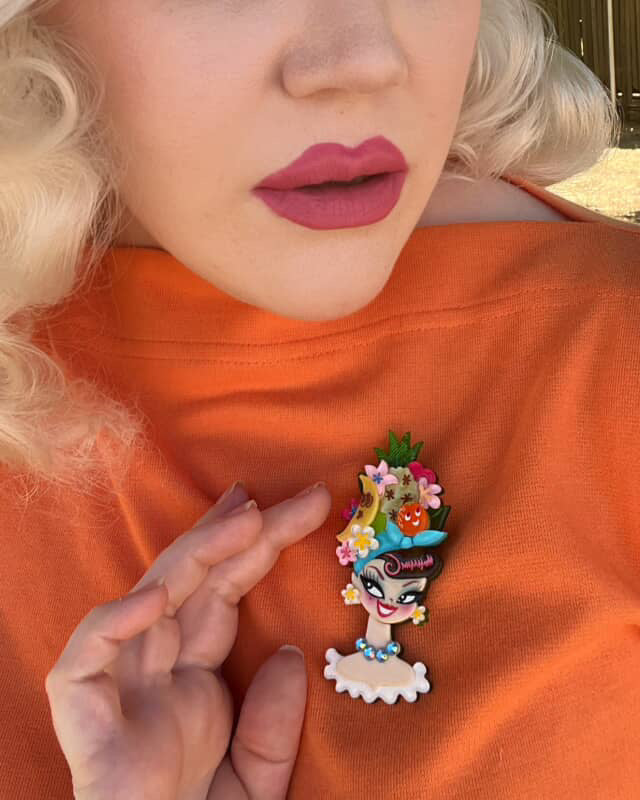 Person wearing an orange shirt with a colorful brooch featuring a cartoon character with flowers on their head.