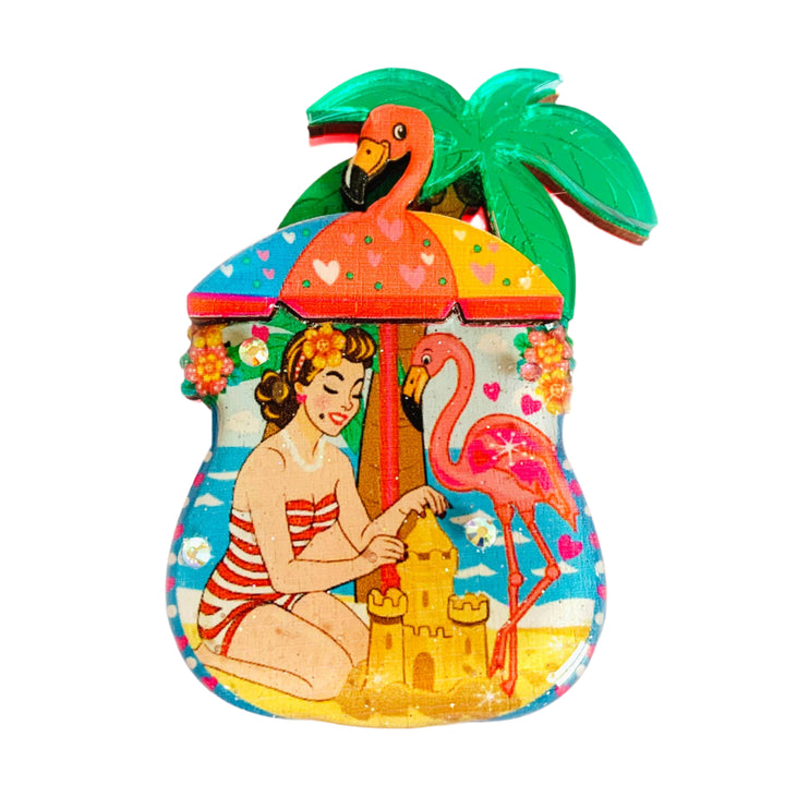 Decorative brooch with a woman building a sandcastle, flamingos, and palm trees on a white background.