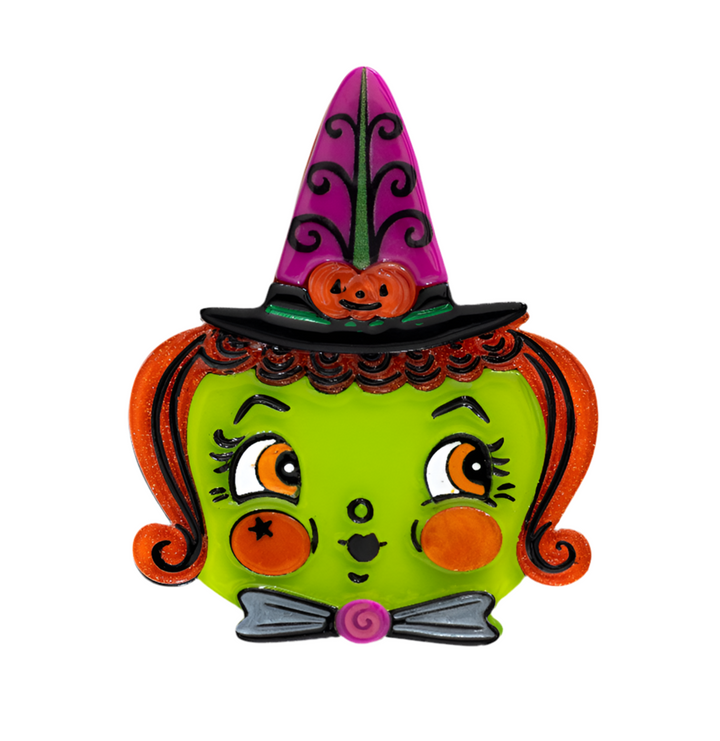 A colorful brooch depicting a whimsical witch character with a green face, orange and black hair, and a purple hat with a pumpkin design.