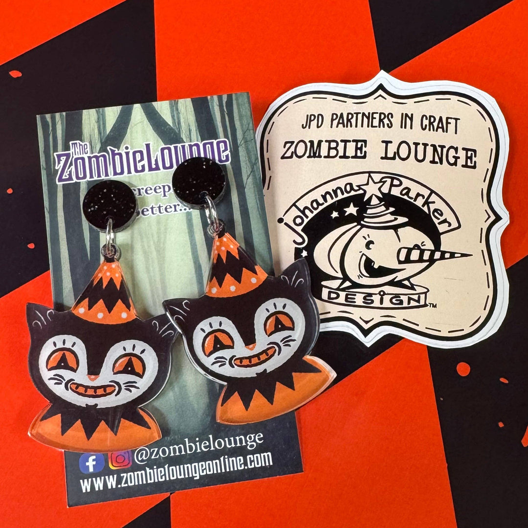 Halloween-themed cat earrings on a Zombie Lounge branded card against an orange and black checkered background.