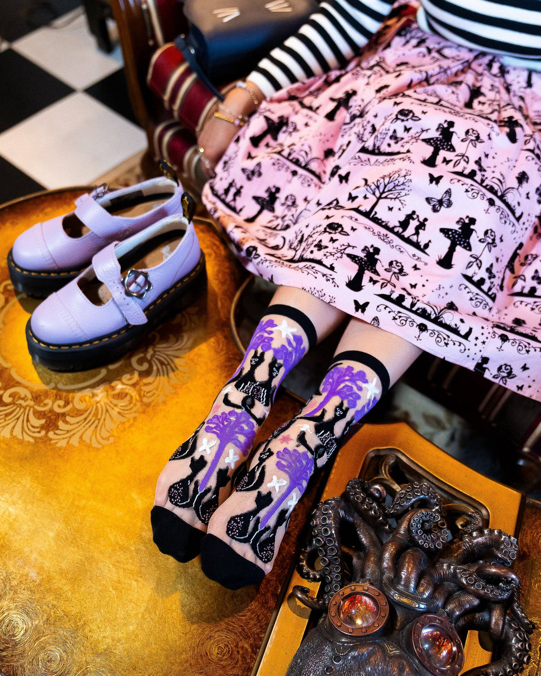 Person wearing purple shoes and socks with black cat design on a checkered floor.