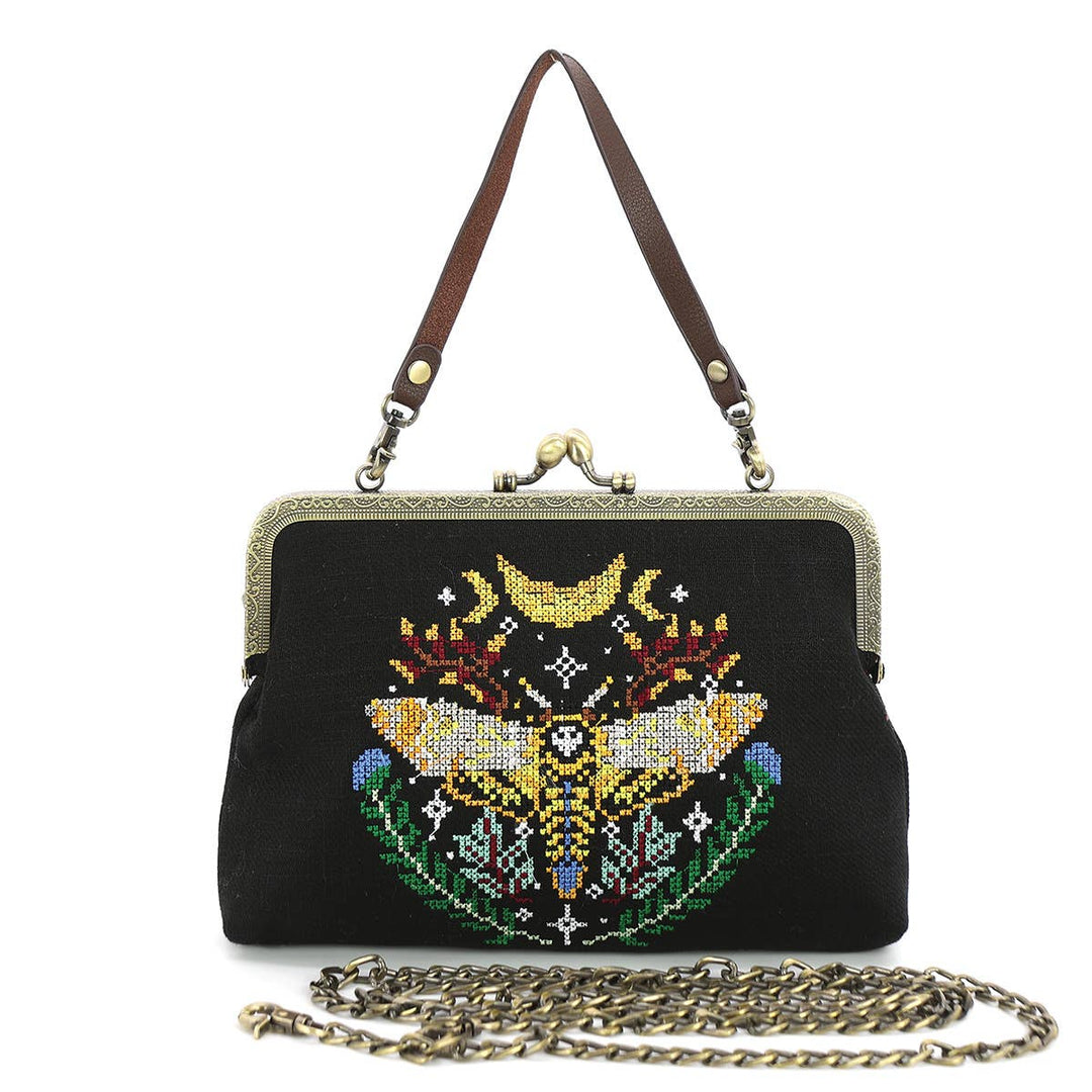 A vintage style handbag with a skull and moth embroidered design on a black canvas background, featuring a metal frame clasp and a leather strap.